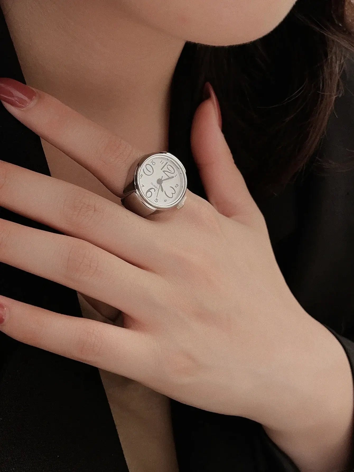 Woman wearing a stylish vintage ring watch with a silver band and classic clock face on her finger against a dark blouse.